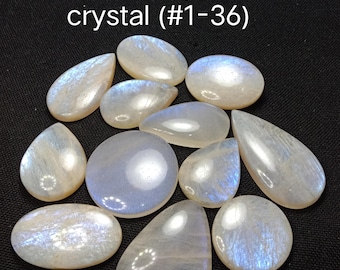 May include: A collection of eleven polished moonstone cabochons in various shapes including oval, round, and teardrop. Each stone displays a translucent, milky white appearance with iridescent blue flashes. The text "Choose your crystal (#1-36)" is visible.