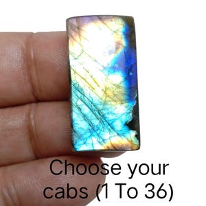 May include: A rectangular labradorite gemstone with a vibrant display of colors, including blues, yellows, and greens. The stone is held in a hand, and the text "Choose your cabs (1 To 36)" is visible.