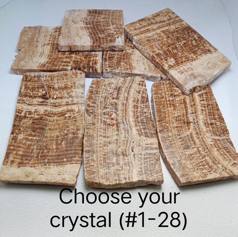 May include: A collection of rectangular brown and beige banded stone slabs with natural layered patterns. The stones feature varied earthy tones and textures. The text "Choose your crystal (#1-28)" is printed in black at the bottom of the image.