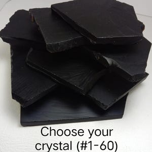 Peut inclure: Une pile de dalles de cristal d'obsidienne noire, plates et irrégulières, aux bords tranchants et dentelés. Les pièces présentent une texture de surface lisse, mate à légèrement réfléchissante. Le texte "Choose your crystal (#1-60)" est imprimé en noir en bas.