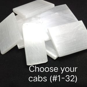 May include: A collection of rectangular white selenite cabochons with a fibrous, translucent texture. The stones are arranged in a pile, and the text "Choose your cabs (#1-32)" is printed in white at the bottom of the image.