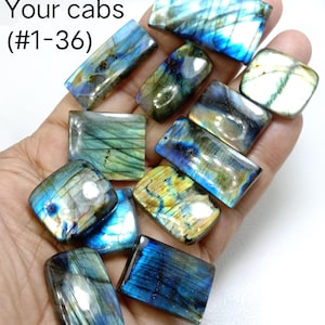May include: A hand holding a collection of polished labradorite cabochons in various rectangular and square shapes. The stones display iridescent blue, green, and yellow flashes. Text in the upper left corner reads "Choose Your cabs (#1-36)".