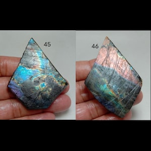 May include: Two iridescent labradorite gemstones, each held in a hand. The stones are roughly diamond-shaped, with a dark base color and flashes of blue, purple, and green. The number 45 is visible on the left stone, and 46 on the right.