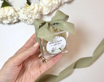 May include: A small, clear, square box with a green ribbon tied in a bow. The box contains a floral design and a white circle with the text "Marco e Giulia 15 Luglio 2023".