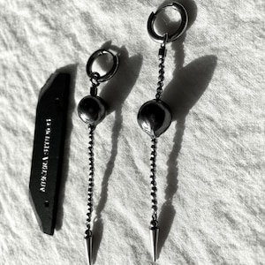 May include: A pair of silver-toned earrings with a dangling beaded chain and a conical spike. Each earring has a circular hoop at the top and a large, round bead. A black, rectangular object with white text is to the left.