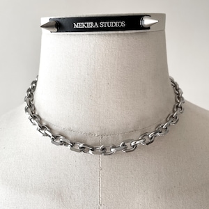 Könnte beinhalten: Eine silberne Halskette und ein schwarzer und silberner Choker mit dem Text "MEKERA STUDIOS" und silbernen Stacheln. Der Schmuck wird auf einer Schaufensterpuppe präsentiert.