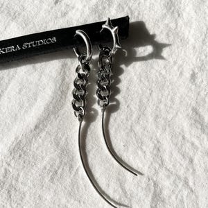 May include: A pair of silver earrings with a chain and a curved spike detail. The earrings are hanging from a black leather background with the text "KERA STUDIOS".