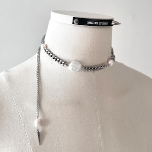 May include: A silver chain necklace with a large, irregular pearl pendant and smaller pearls. A black choker with a silver spike and the text "MEKERA STUDIOS" is also visible. The necklace is displayed on a mannequin.