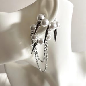 May include: A unique earring design featuring a cluster of white faux pearls and silver-toned spikes. The earring has dangling chains and is displayed on a white mannequin ear. The jewelry has a modern, edgy aesthetic.