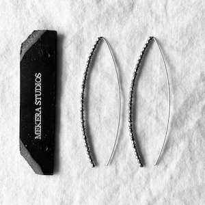 May include: A pair of silver-toned curved wire earrings featuring a beaded texture along the outer edge. Beside the earrings is a rectangular black leather tag with the white text MEKERA STUDIOS printed vertically on the surface.