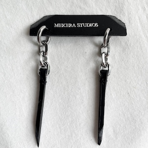 May include: A pair of black leather earrings with silver chain and hoop accents. The earrings are displayed on a black card with the text "MEKER STUDIOS".