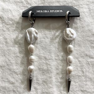 May include: A pair of dangling earrings with white pearls and silver-tone metal accents. The earrings have a large, irregular pearl at the top, followed by smaller pearls, and a silver spike at the bottom. The earrings are displayed on a black tag with the text "MEKERA STUDIOS".