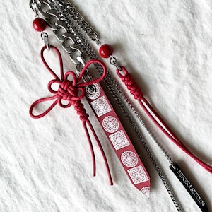 May include: A red and silver keychain with multiple chains, red beads, and a red decorative knot. A red tag with a white geometric pattern is attached. A black tag with the text "MEKERA STUDIOS" is also attached.