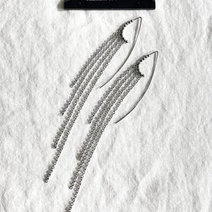 May include: A pair of silver chain earrings with a cascading design. Each earring features a curved hook and multiple strands of delicate chains. The earrings are displayed on a white fabric surface, with a black tag that reads "MEKERA STUDIOS".
