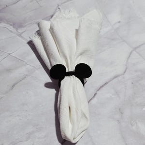 Könnte beinhalten: Eine weiße Stoffserviette, gehalten von einem schwarzen Mickey Mouse Ohren-Serviettenring. Die Serviette ist gefaltet und hat eine strukturierte Oberfläche. Der Serviettenring ist einfarbig, matt schwarz.
