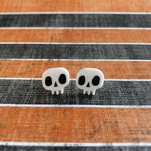 May include: A pair of white skull-shaped stud earrings with black eye and nose sockets. The earrings are displayed on a background with orange, black, and gray horizontal stripes. These earrings are a spooky accessory.