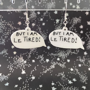 May include: White speech bubble earrings with the text "BUT I AM LE TIRED!" in black. The earrings hang from silver-colored hooks. The background is black with white star and dot patterns.
