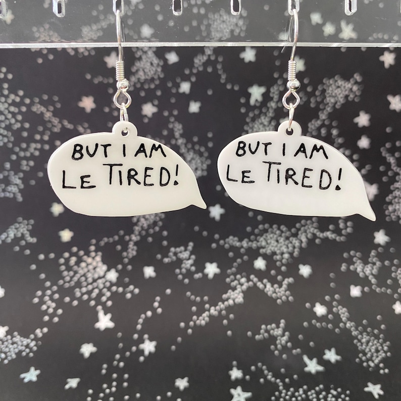 May include: White speech bubble earrings with the text "BUT I AM LE TIRED!" in black. The earrings hang from silver-colored hooks. The background is black with white star and dot patterns.