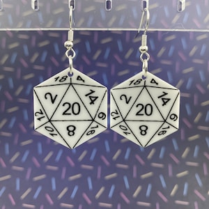 May include: A pair of white dangle earrings shaped like a twenty-sided die, also known as a d20. The earrings have black numbers and lines on them.