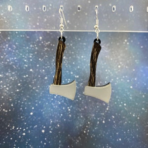 May include: A pair of silver axe earrings with a wood-grain design. The earrings are hanging from a silver hook.