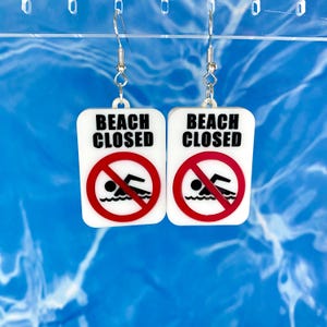 May include: A pair of white earrings shaped like a beach closed sign with a red circle and a no swimming symbol. The sign says "Beach Closed" in black text.