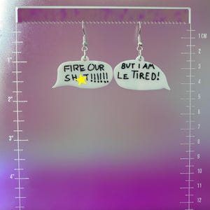May include: White acrylic earrings in the shape of speech bubbles. One earring reads "FIRE OUR SH T!!!!!!" with a yellow star, and the other says "BUT I AM LE TIRED!" Silver-colored fish hook ear wires.