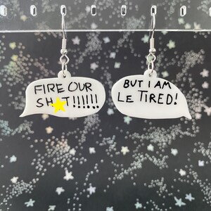 May include: A pair of white acrylic earrings in the shape of speech bubbles. One earring reads "FIRE OUR SH T!!!!!!" with a yellow star, and the other says "BUT I AM LE TIRED!" in black lettering. The earrings hang from silver-colored hooks.