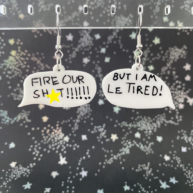 May include: A pair of white acrylic earrings in the shape of speech bubbles. One earring reads "FIRE OUR SH T!!!!!!" with a yellow star, and the other says "BUT I AM LE TIRED!" in black lettering. The earrings hang from silver-colored hooks.