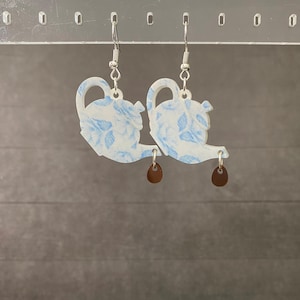 May include: A pair of dangle earrings featuring a teapot design. The earrings are white with a blue floral pattern and a small brown teardrop charm. The earrings hang from silver-colored hooks.