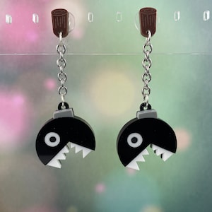 May include: A pair of black and white earrings featuring a video game character with a large mouth and a single eye. The earrings are attached to silver chains and have a brown top.