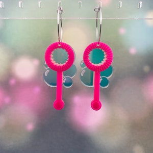 May include: A pair of pink bubble wand earrings with silver hoops. The bubble wands have a pink handle and a round, pink bubble blower.