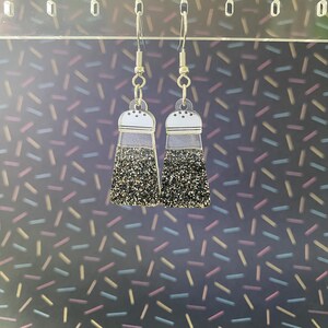 Puede incluir: Un par de pendientes originales con forma de saleros. Los pendientes tienen una parte superior transparente con un borde blanco y una base con purpurina negra. Cuelgan de ganchos para las orejas de color plateado.