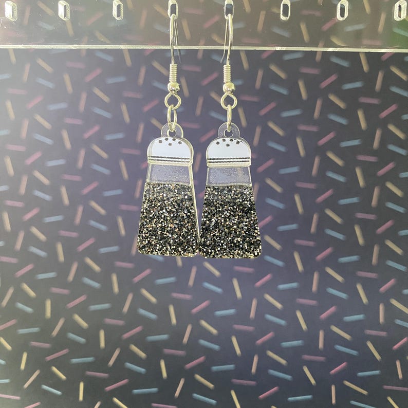 Puede incluir: Un par de pendientes originales con forma de saleros. Los pendientes tienen una parte superior transparente con un borde blanco y una base con purpurina negra. Cuelgan de ganchos para las orejas de color plateado.