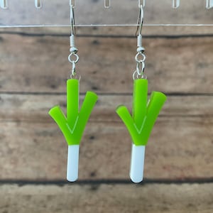 May include: A pair of dangling earrings featuring a leek design. Each earring has a bright green top and a white base, with silver-colored hooks. The earrings are displayed against a wooden background.
