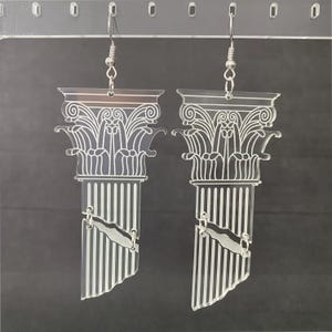 May include: A pair of clear acrylic drop earrings shaped like broken classical Greek columns. Each earring features a detailed white etched design of a Corinthian capital and a fluted shaft, suspended from silver-toned metal fishhook ear wires.