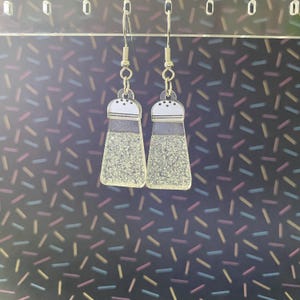 Puede incluir: Pendientes novedosos con forma de saleros. Los pendientes transparentes tienen un interior brillante, una parte superior blanca con puntos negros y una banda gris. Cuelgan de ganchos para las orejas de color plateado.