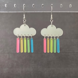 May include: A pair of cloud-shaped earrings with colorful rain drops. The earrings feature a white cloud design with silver hooks. Each cloud has six colorful, rectangular rain drops in pink, yellow, green, and blue.