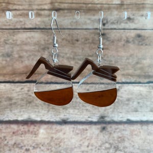 May include: A pair of coffee pot shaped earrings with a clear resin body and brown coffee inside. The earrings are silver and have a hook closure.