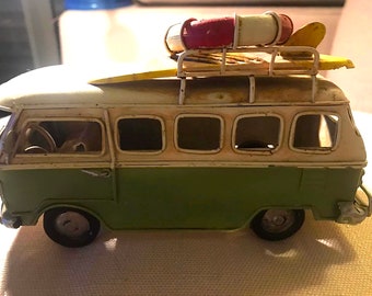 May include: A green and white toy van with a surfboard and a red and white life preserver on the roof. The van has a white roof and a green body with a white stripe. The van has four windows on each side.