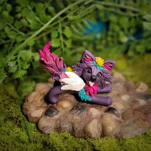 May include: A purple and pink clay figurine of a wolf with a white wing and a flower crown. The wolf is sitting on a brown rock with moss around it.