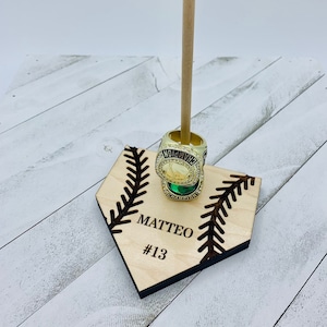 May include: A wooden baseball home plate stand with a baseball stitch design. The stand has a wooden dowel for holding rings. The stand is personalized with the name "MATTEO" and the number "#13".