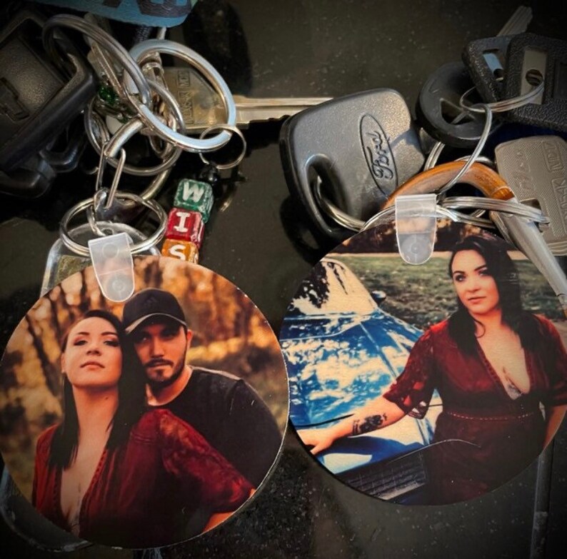 May include: Two round keychains with photos printed on them. One keychain features a couple in a wooded area, the other features a woman standing next to a blue car.