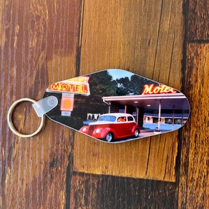 May include: A red and white vintage car parked in front of a motel with a neon sign that reads "Motel". The car is a classic 1940s or 1950s model with a chrome bumper and whitewall tires. The motel sign is lit up with bright neon lights. The keychain is made of plastic and has a metal ring.