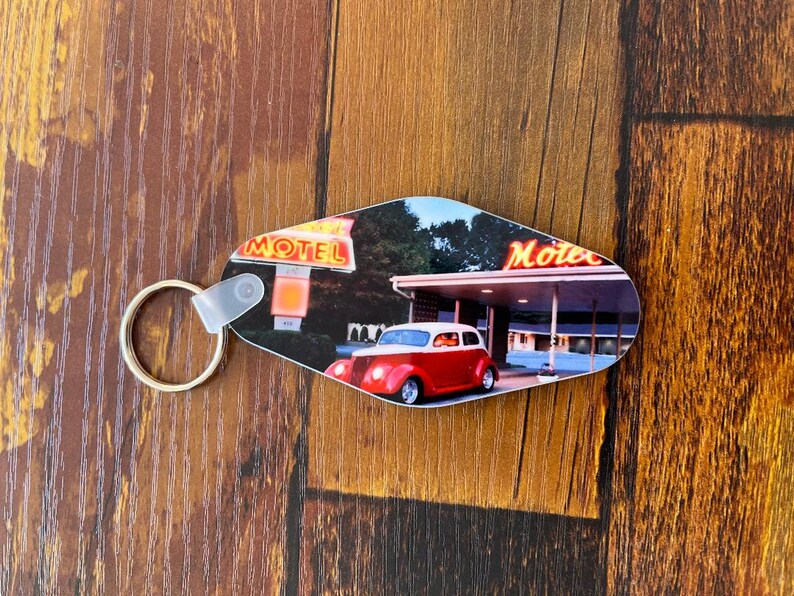 May include: A red and white vintage car parked in front of a motel with a neon sign that reads "Motel". The car is a classic 1940s or 1950s model with a chrome bumper and whitewall tires. The motel sign is lit up with bright neon lights. The keychain is made of plastic and has a metal ring.