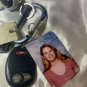 May include: A keychain with a blue and silver carabiner, a black key fob with the words "lock" and "unlock", and a small white figurine with a black dot. The keychain also has a photo card with a picture of a young woman with long blonde hair wearing a pink and white polka dot shirt.