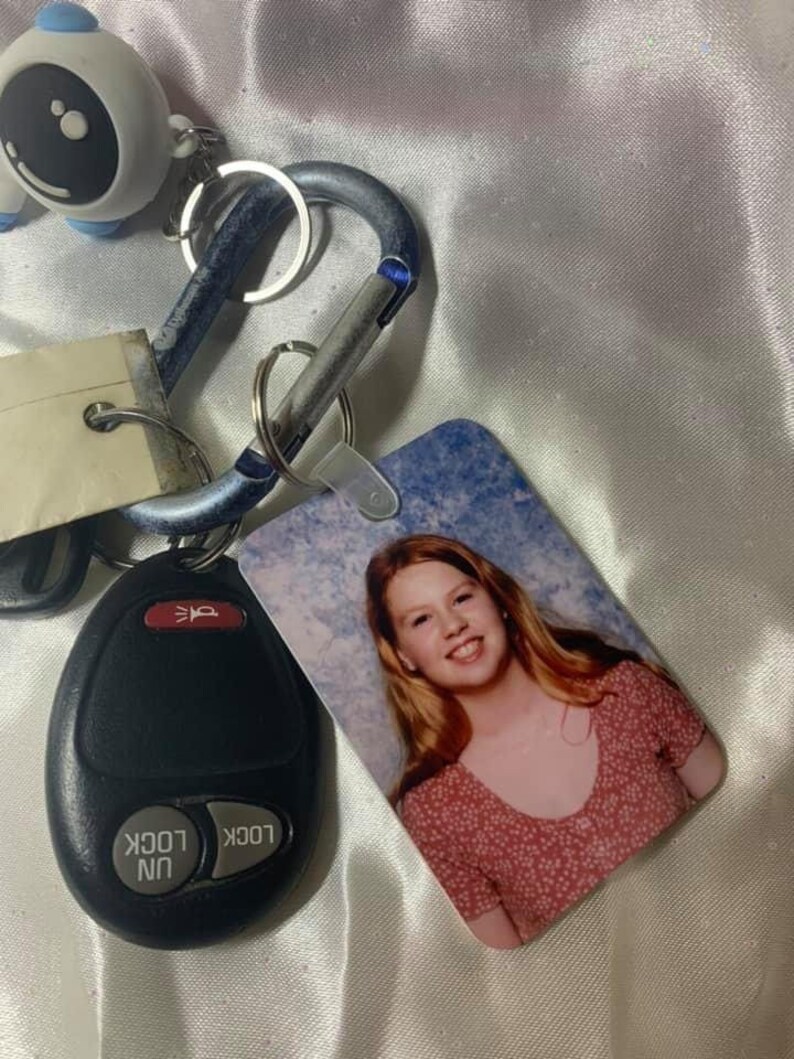 May include: A keychain with a blue and silver carabiner, a black key fob with the words "lock" and "unlock", and a small white figurine with a black dot. The keychain also has a photo card with a picture of a young woman with long blonde hair wearing a pink and white polka dot shirt.