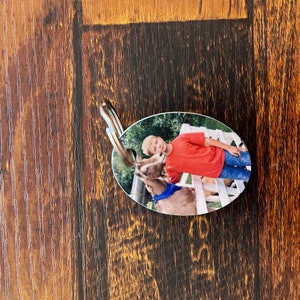 May include: Oval-shaped keychain with a photo of a young person in a red shirt and blue jeans standing next to a brown and white goat. The goat is wearing a blue bandana.