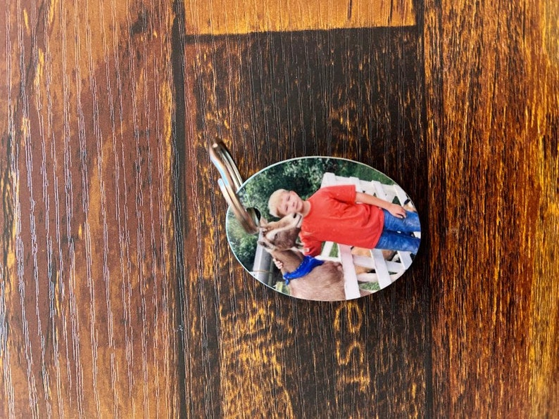 May include: Oval-shaped keychain with a photo of a young person in a red shirt and blue jeans standing next to a brown and white goat. The goat is wearing a blue bandana.