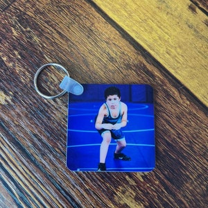 May include: A blue and white wrestling singlet keychain with a photo of a young wrestler in a wrestling stance on a blue mat.