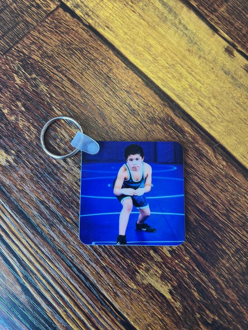 May include: A blue and white wrestling singlet keychain with a photo of a young wrestler in a wrestling stance on a blue mat.